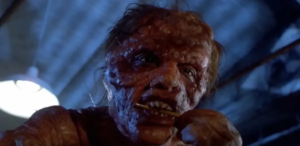 Primer plano de una criatura humana deforme masticando un objeto metálico, escena de transformación corporal en la película La mosca.