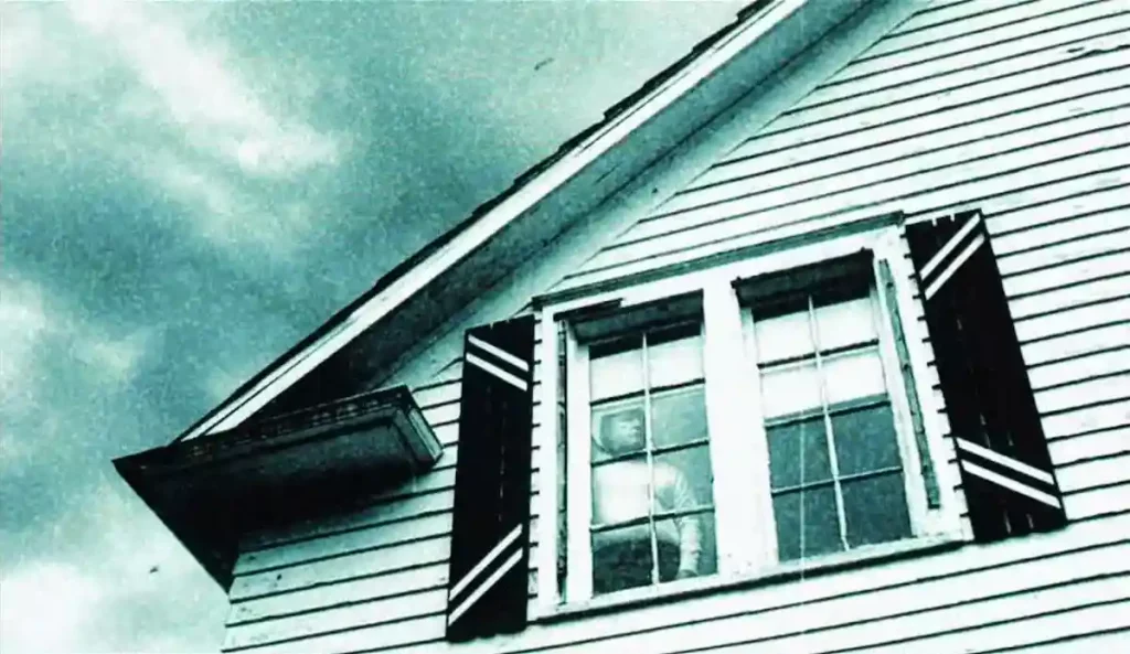 Plano de una casa blanca con cielo nublado, donde un hombre aparece observando desde una ventana del piso superior.