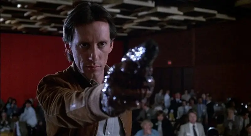 Hombre con una pistola orgánica en lugar de mano, apuntando con determinación hacia el frente, escena de la película Videodrome.