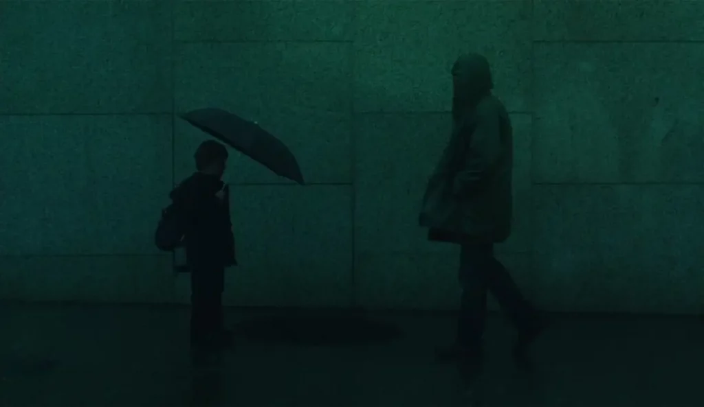 Niño sosteniendo un paraguas bajo la lluvia frente a un hombre encapuchado, en una escena oscura y minimalista.