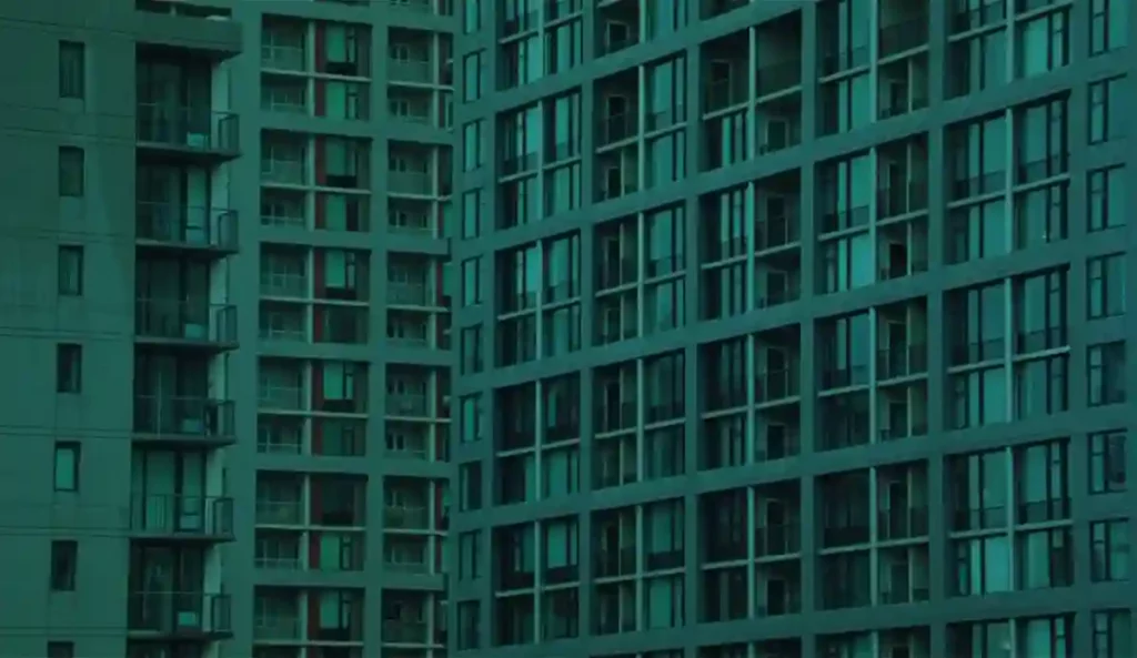 Fachada repetitiva de varios bloques de pisos, con decenas de ventanas iluminadas al anochecer.