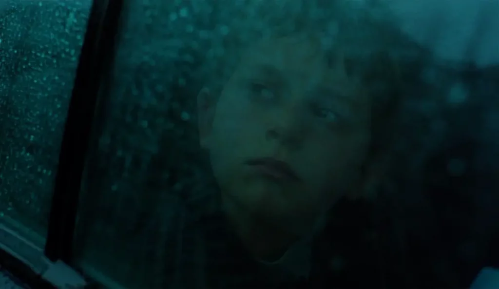 Aidan mira con expresión melancólica a través del cristal de un coche cubierto de gotas de lluvia.