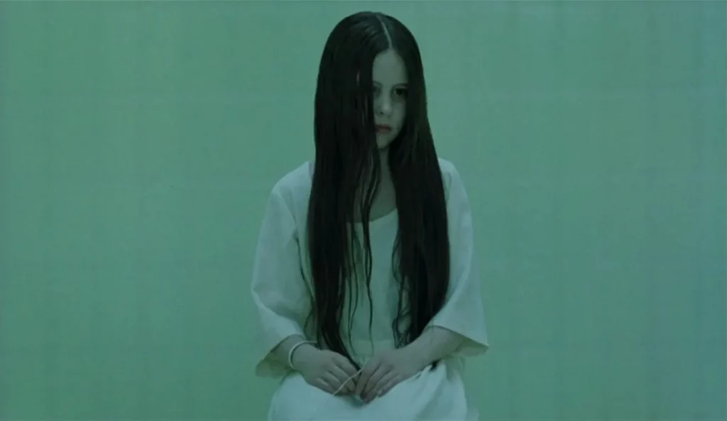 Samara Morgan sentada en una habitación esterilizada, con el cabello largo cubriéndole parte del rostro y expresión ausente.