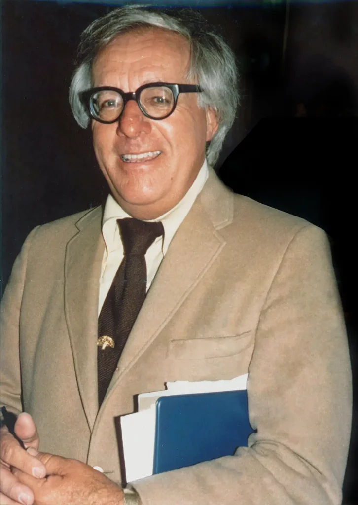 Escritor Ray Bradbury sonriendo, vestido con traje claro y corbata oscura, sosteniendo libros y un bolígrafo.