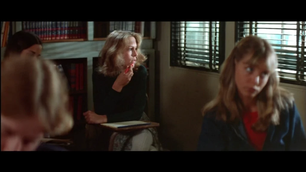 oven en clase mira por la ventana mientras continúa la lección; persianas y estanterías al fondo.