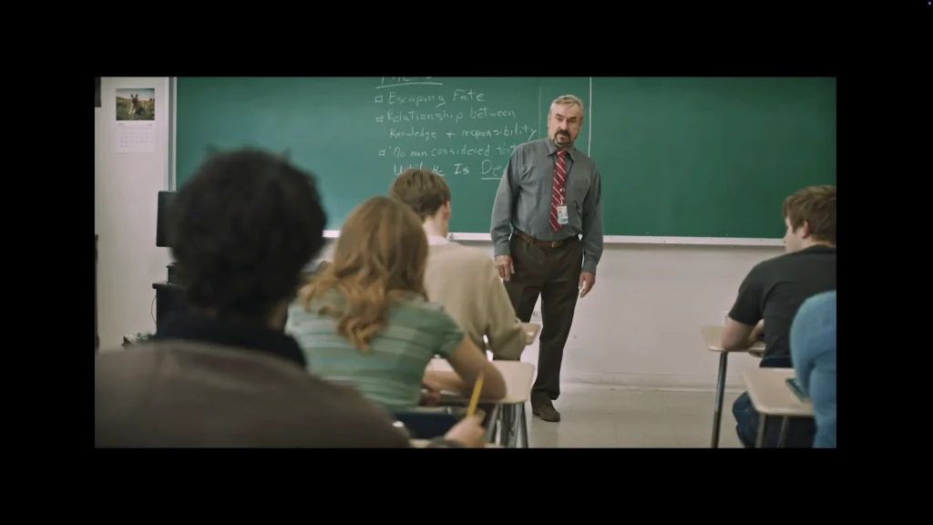 Profesor frente a una pizarra verde hablando a una clase; alumnos de espaldas, lista de temas escrita en la pizarra.