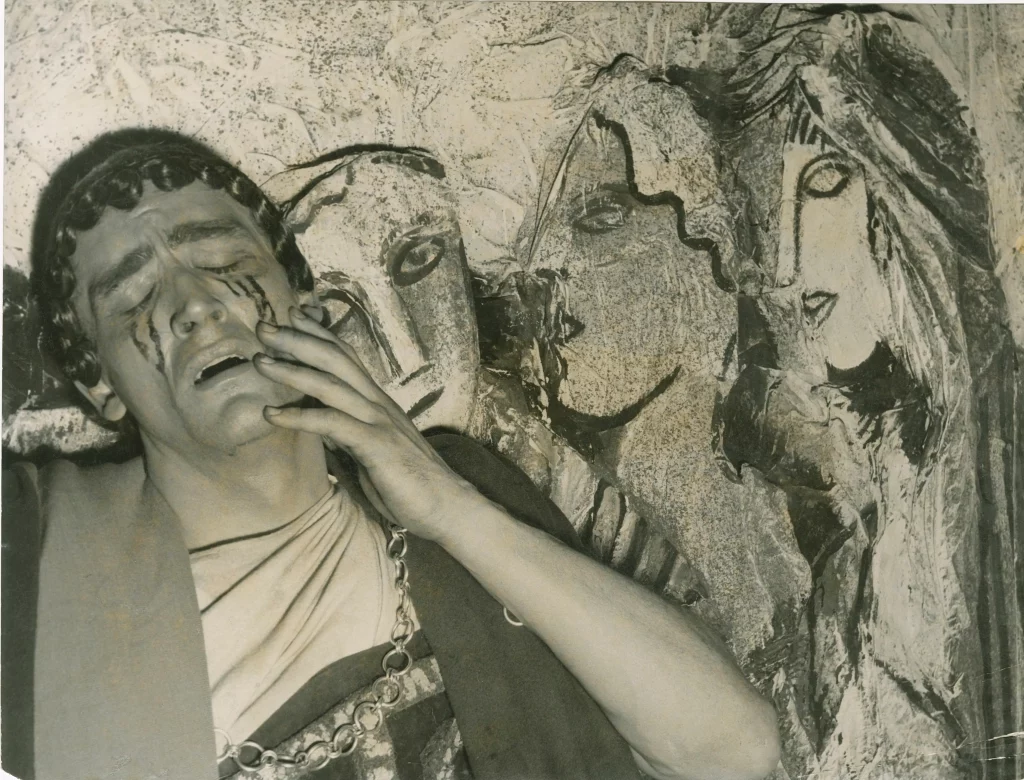 Actor de tragedia griega en escena, rostro con lágrimas pintadas junto a máscaras del coro, foto en blanco y negro.