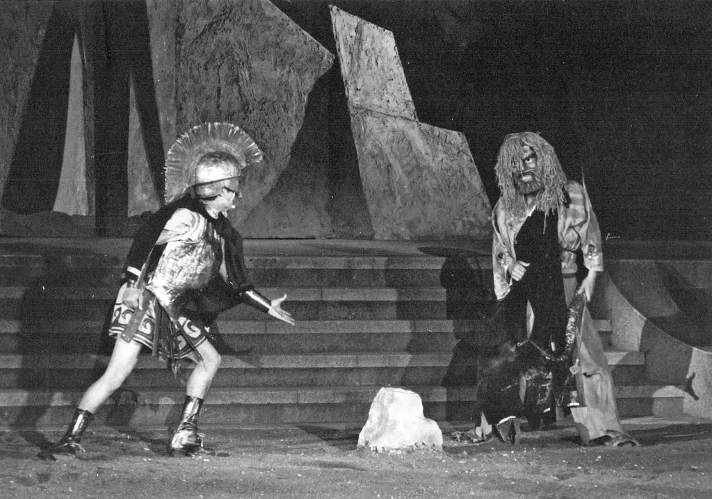 Escena teatral de tragedia clásica: actor con armadura se encara a figura barbada; escenografía geométrica, fotografía de 1962.