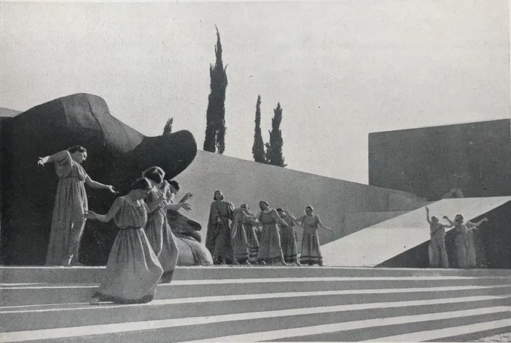 Coro de actores con túnicas desciende una gran escalinata exterior; escenografía geométrica en blanco y negro.