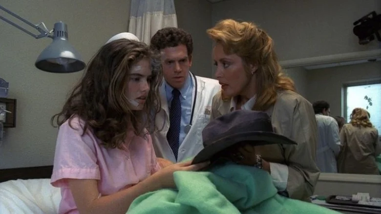 Nancy en una cama de hospital sostiene un sombrero oscuro mientras un médico y su madre observan con preocupación.