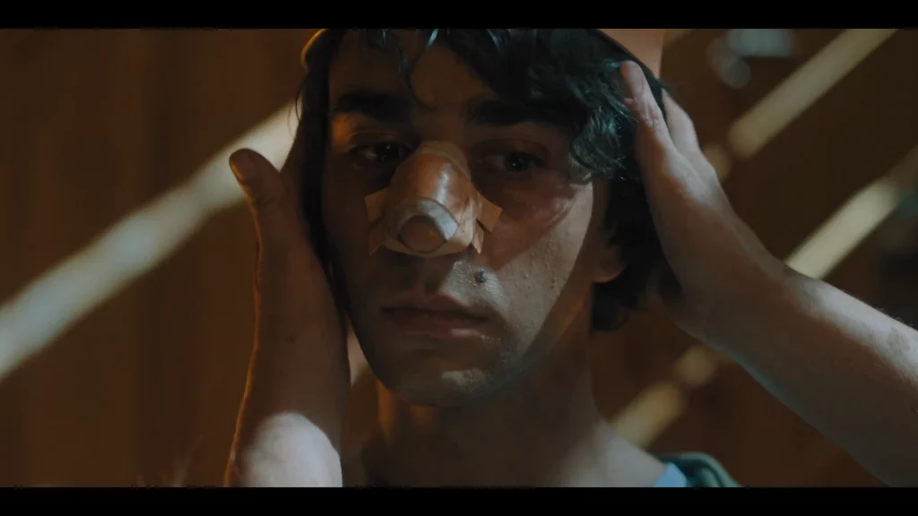 Coronación ritual de Peter: dos manos le ciñen la cabeza mientras mira fijo, luz cálida en penumbra.