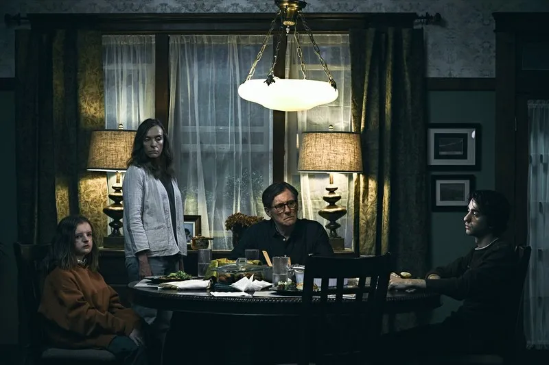 La familia Graham sentada alrededor de la mesa, miradas tensas bajo una lámpara colgante; ambiente oscuro.