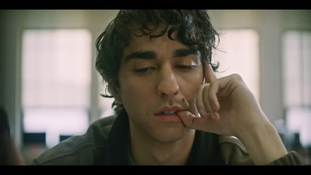 Primer plano de Peter en clase, con la mirada baja y el dedo en los labios, luz de ventana desenfocada al fondo.