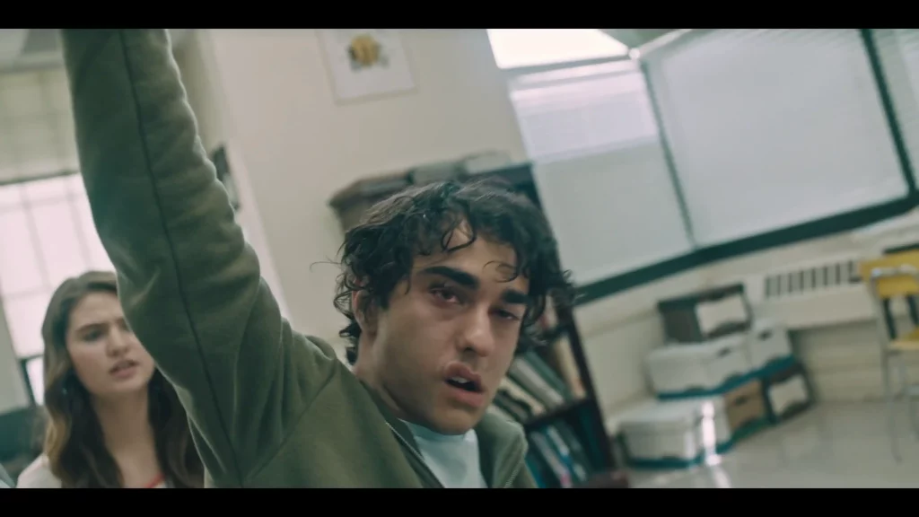 Primer plano de un adolescente en clase con el brazo alzado de forma rígida y mirada aturdida; aula luminosa y estanterías al fondo.