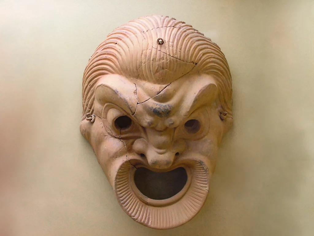 Máscara teatral griega de terracota con ceño fruncido y gran boca abierta, vista frontal sobre fondo neutro.