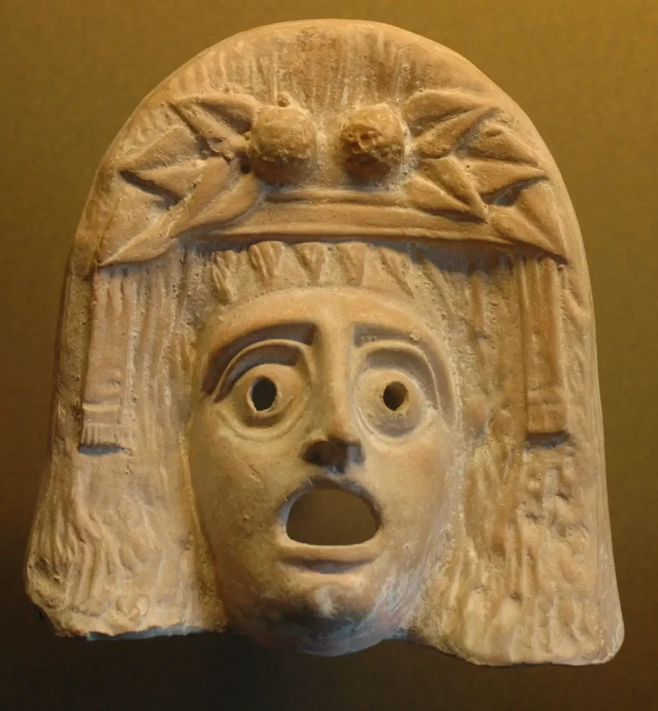 Primer plano de una máscara teatral de Dionisos en terracota, con ojos perforados y boca abierta, expuesta en el Louvre.